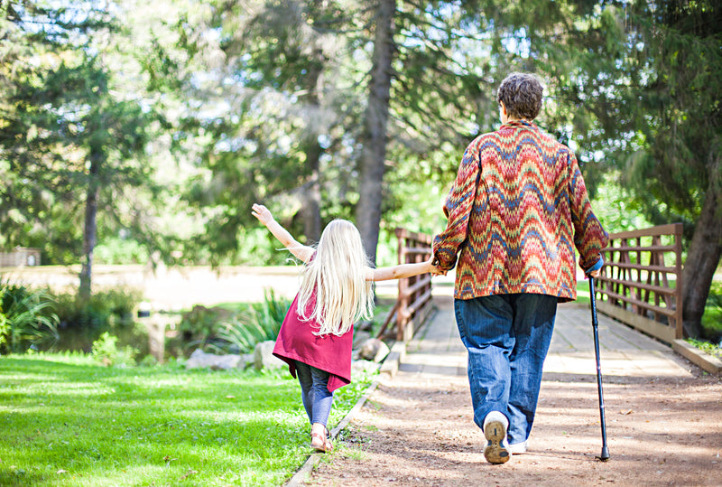 Woman with a cane walking hand-in-hand with a young girl on a path in a park.