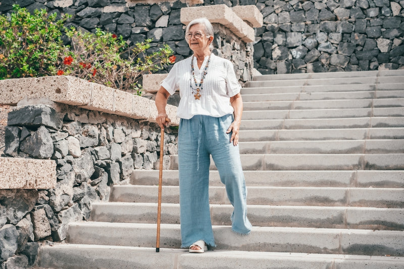 Woman with a walking stick standing on stone steps with a stone wall and plants in the background